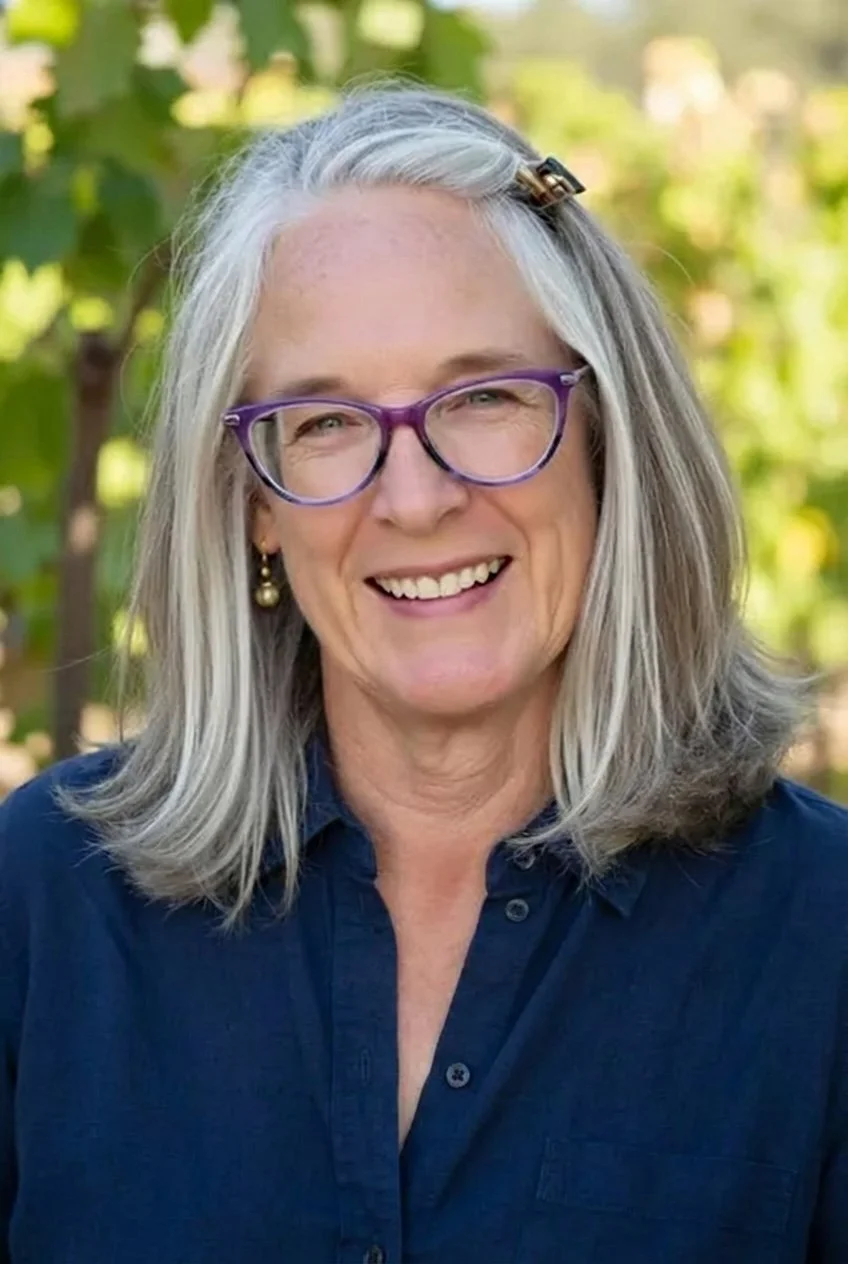 Dawn Dolan close up AI Headshot by Purpose and Pixel, LLC. Dawn is wearing a navy blue button down shirt, purple framed eyeglasses, gold pearl drop earrings and her gray shoulder length hair is worn loose with one small clip on the right side.