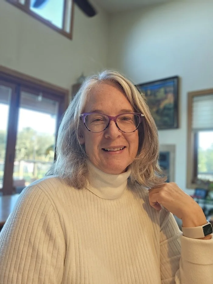 Dawn Dolan's 'before' selfie for her AI photoshoot. She is wearing a white ribbed turtleneck sweater, eyeglasses with purple frames, and her shoulder length gray hair is worn loose. She is smiling at the camera