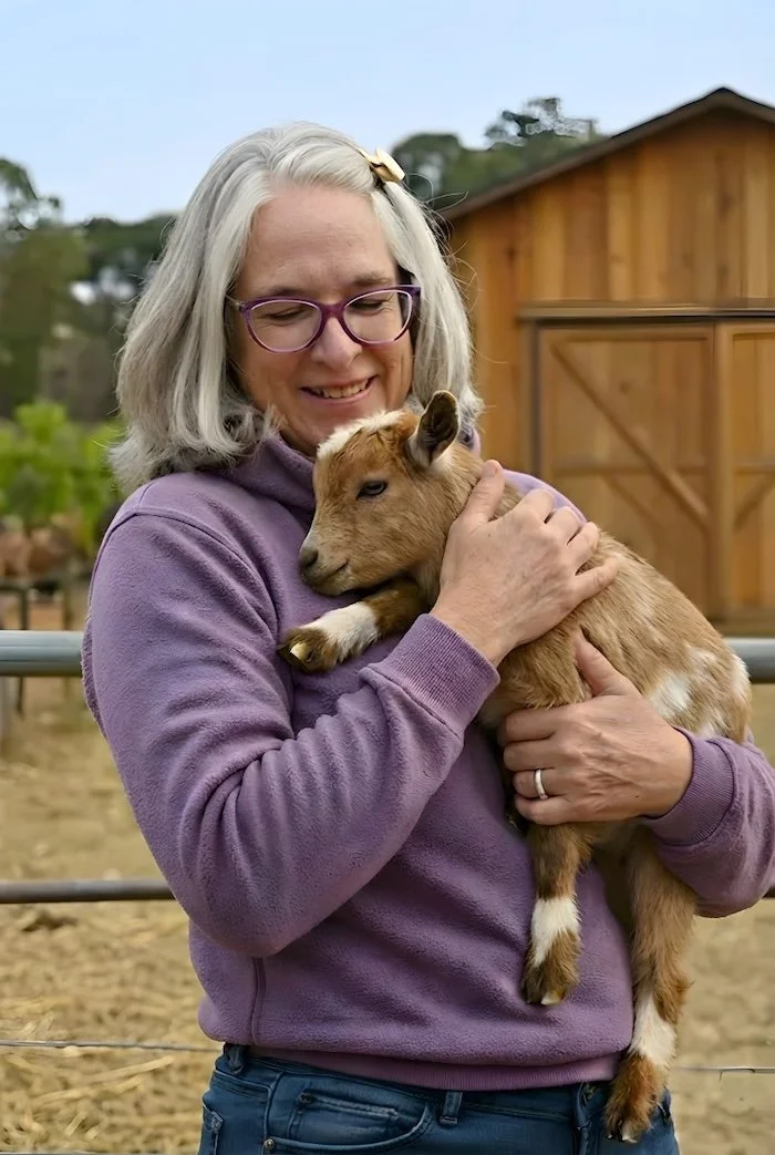 Dawn Dolan AI Headshot by Purpose & Pixel, LLC She is standing in front of her barn on the vineyard wearing a purple fleece top and jeans. She is cradling a brown baby nigerian dwarf goat in her arms and gazing down upon it.