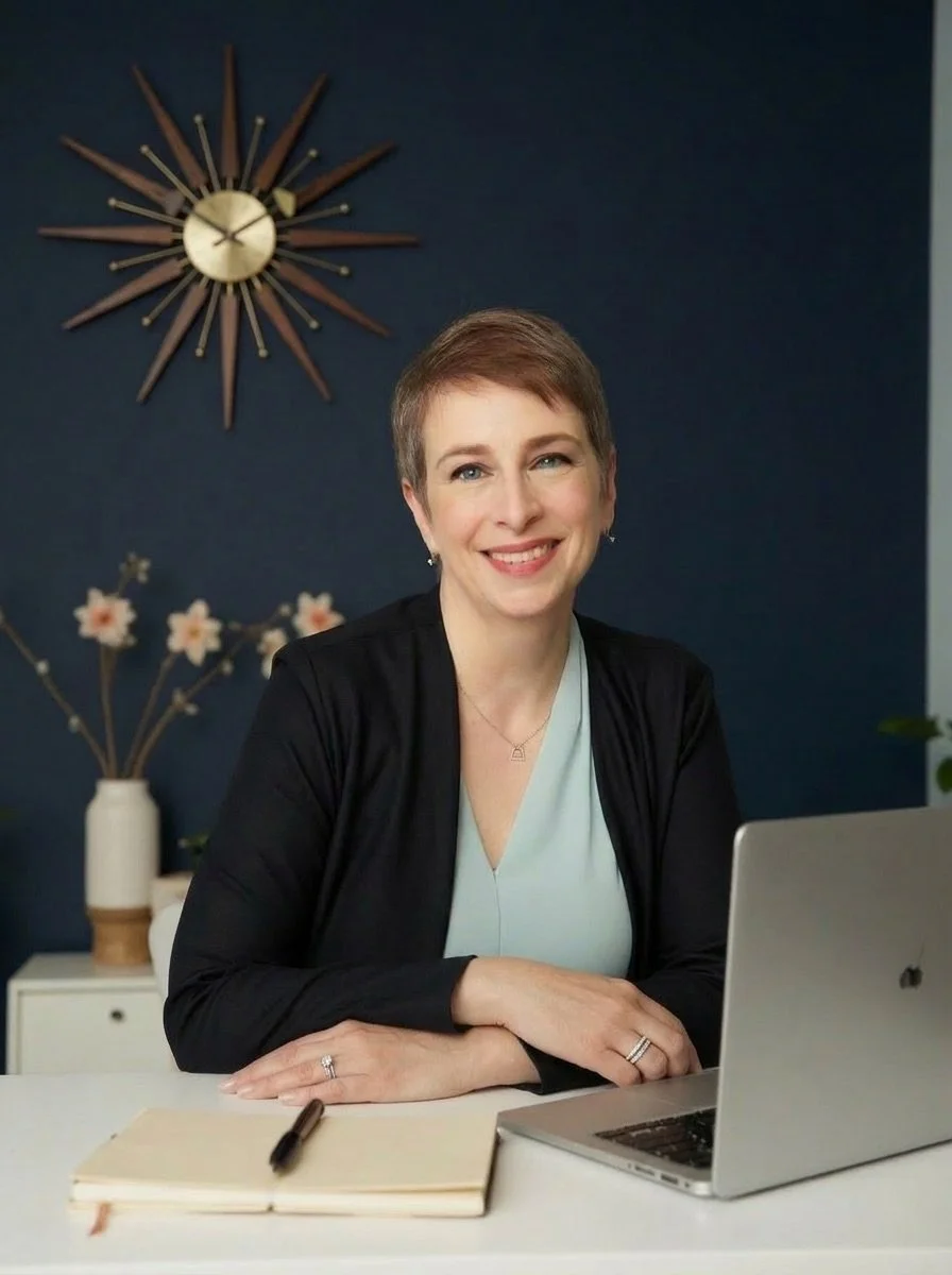 Meg Desjarlais, founder of Purpose & Pixel, seated at a desk with a laptop and notebook, wearing a black blazer, navy blue wall with mid-century sunburst clock in background