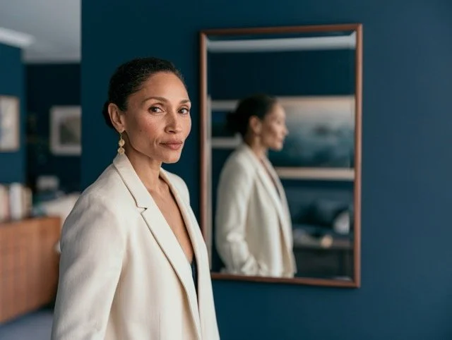 A confident professional woman in a cream blazer stands before a navy wall, her reflection visible but soft in a walnut-framed mirror behind her.