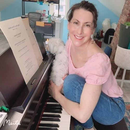 Ilana Davidson, Professional Soprano, sitting at her piano with her small white dog, she is wearing pink shirt and blue jeans, her dark her is in a bun and she is looking at the camera and smiling