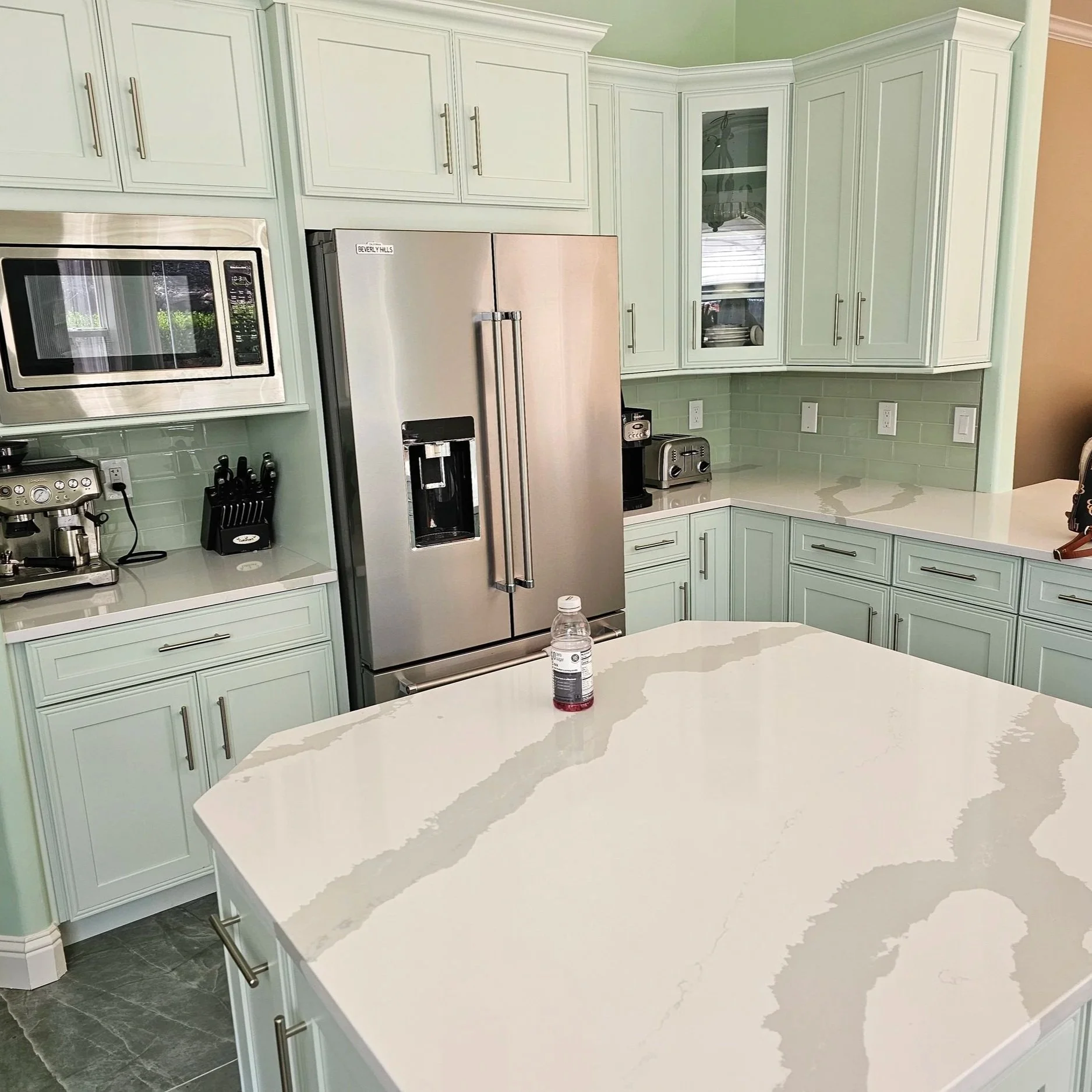 A modern kitchen featuring white cabinetry, a stainless steel refrigerator, a microwave, and a white marble countertop island with gray veining. The kitchen has pastel green walls, a light green backsplash with subway tiles, and black kitchen accessories.