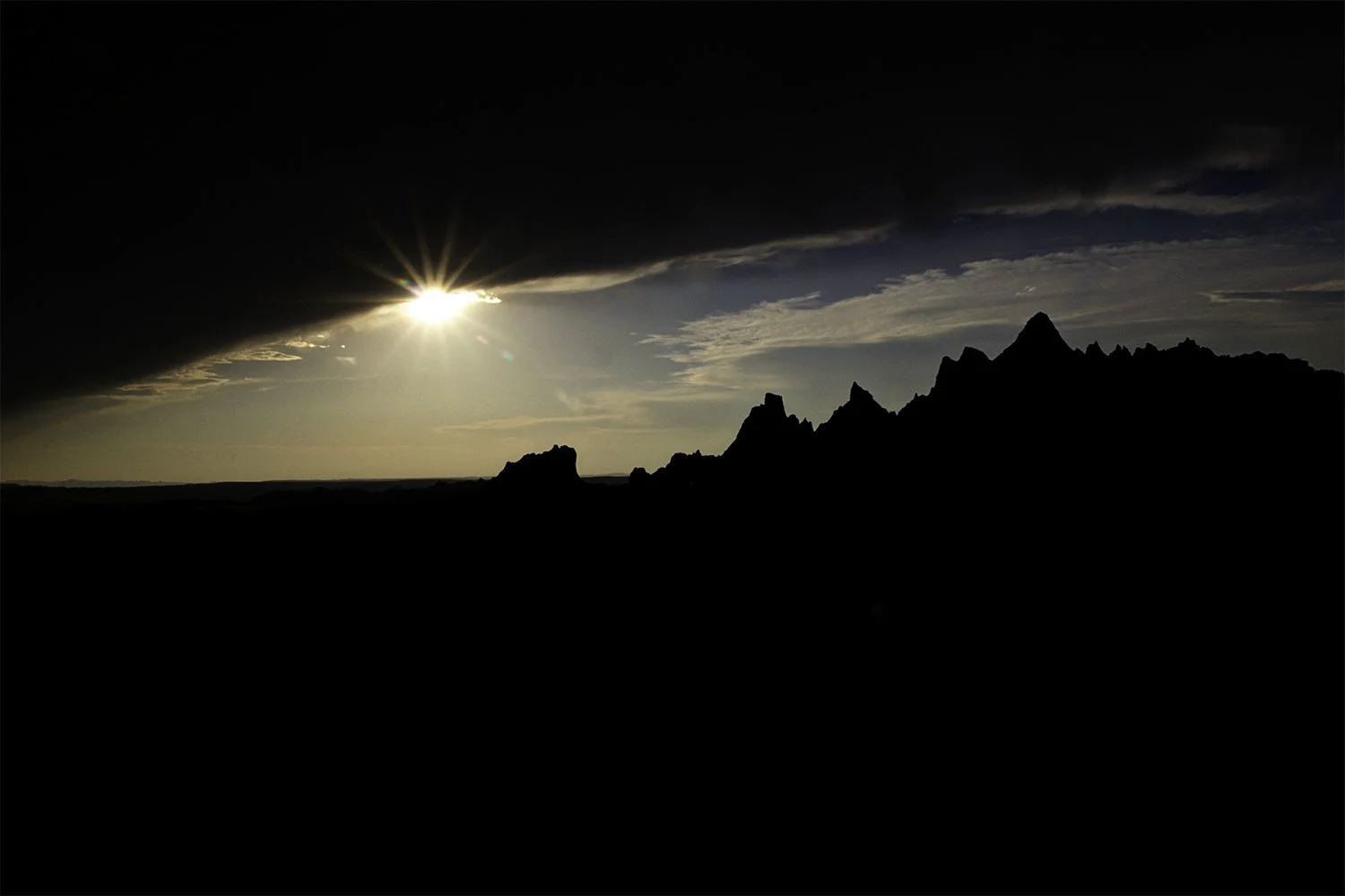 Sunset in Badlands National Park South Dakota.   Photo is framed by the landscape in shadow and a large cloud at the top. Sweeping through the frame is a peek at the sky with the sun’s rays shining through. 