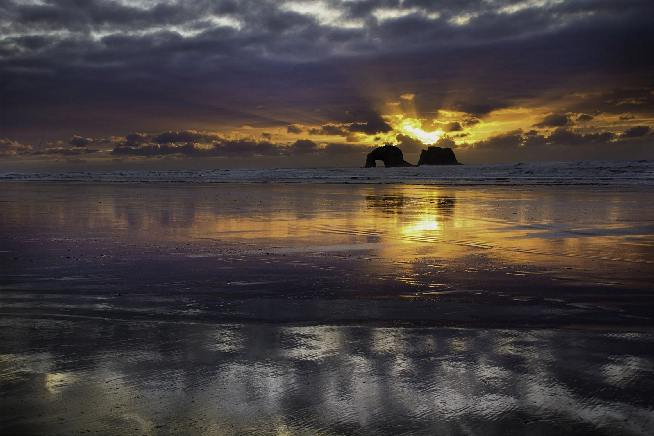 Rockaway Beach Oregon Twin Rocks at sunset. Sun rays behind the rocks, sunset reflecting in the ocean and wet sand. 