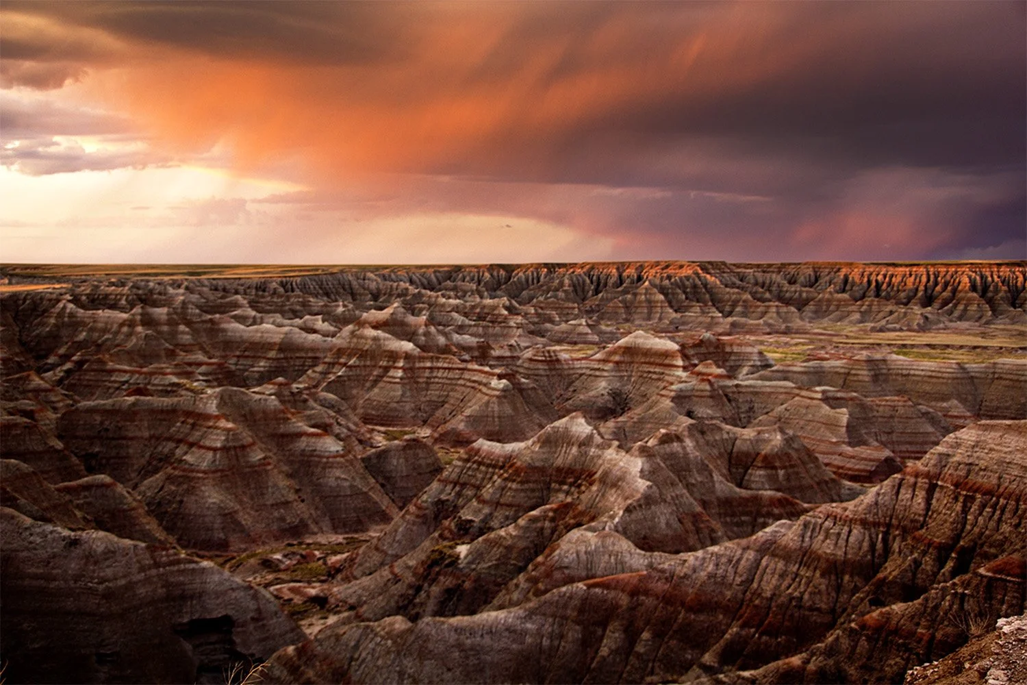 View of the Badlands at Sunset. Picture has red hues and there’s a storm to the right. 
