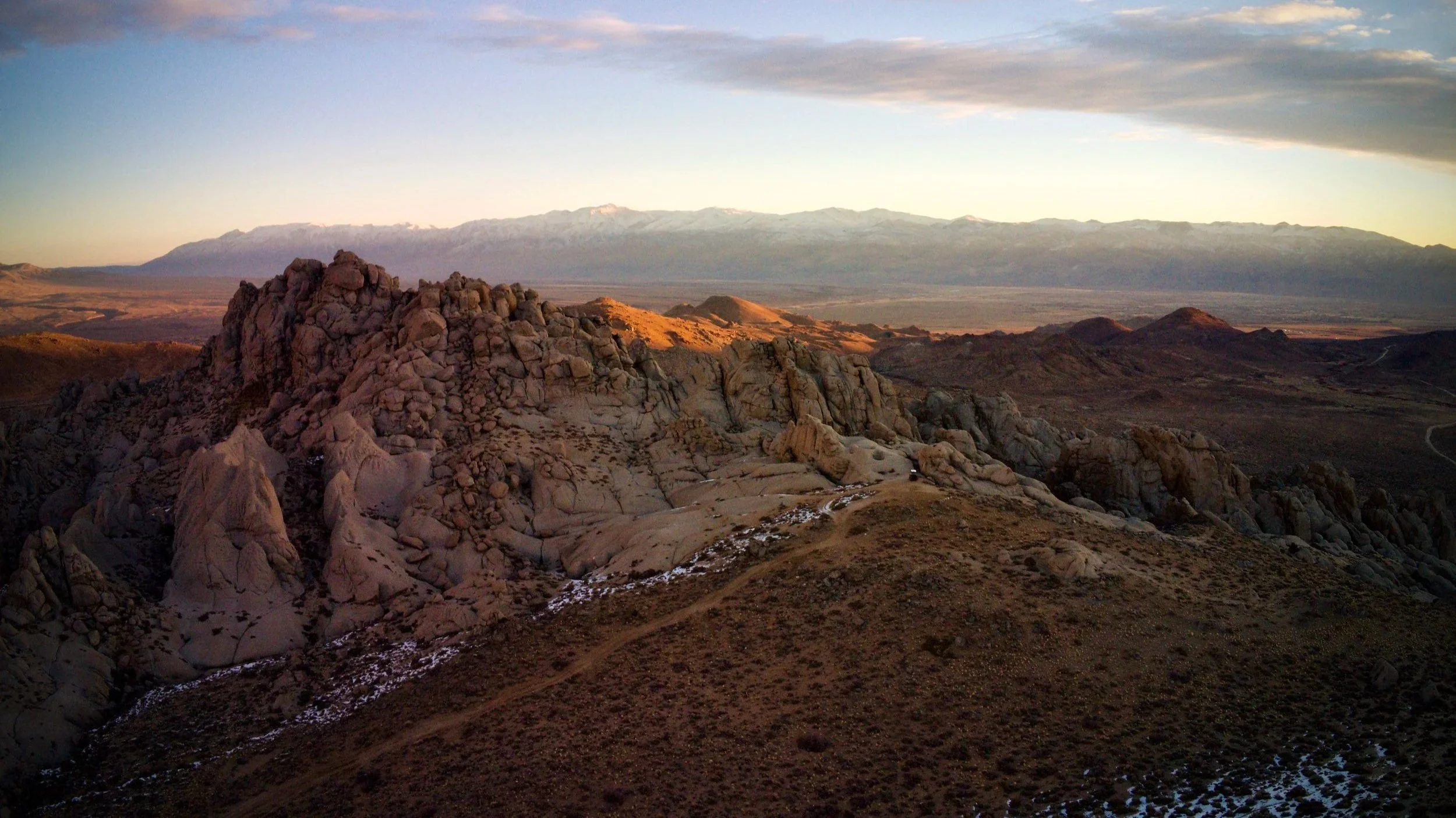 Mountain landscape during sunset with rocky terrain in the foreground, plains, and snow-capped mountains in the background under a partly cloudy sky.