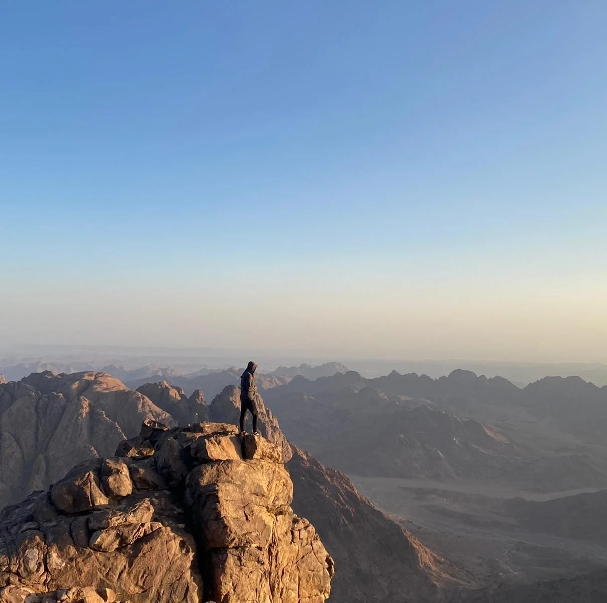 Person standing on rocky mountain peak overlooking a vast desert landscape during sunrise or sunset.