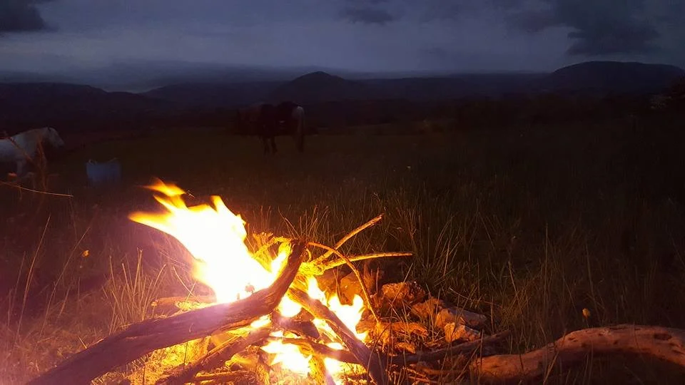 A campfire on a grassy field during dusk or night with dark hills or mountains in the background.