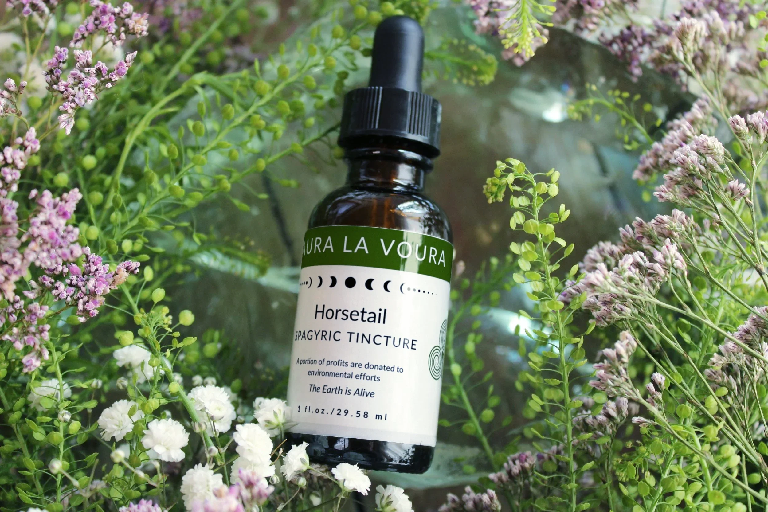 A dark glass dropper bottle labeled "Laura La Voura" with the product name "Horsetail Spagyric Tincture" surrounded by purple and white flowers and green leaves.