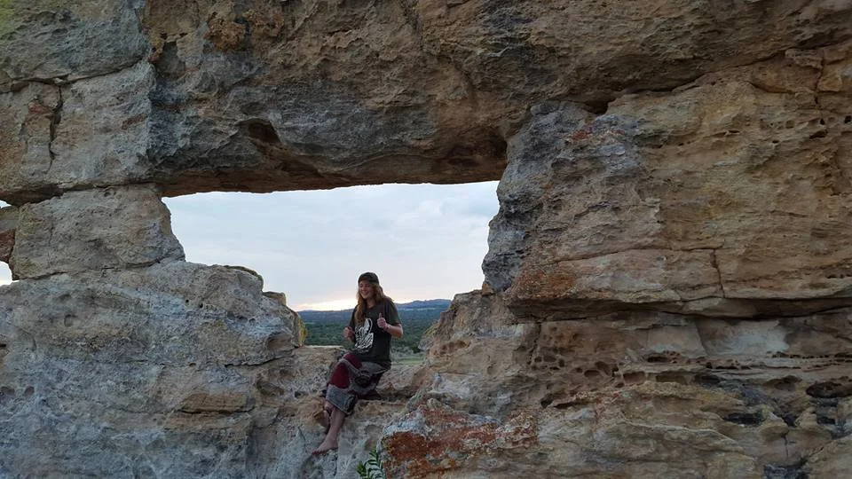A woman sitting on a large rock formation with a natural window, overlooking a landscape with hills and trees during sunset.