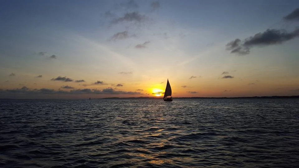 A sailboat on calm water during sunset with a mostly clear sky and some clouds.