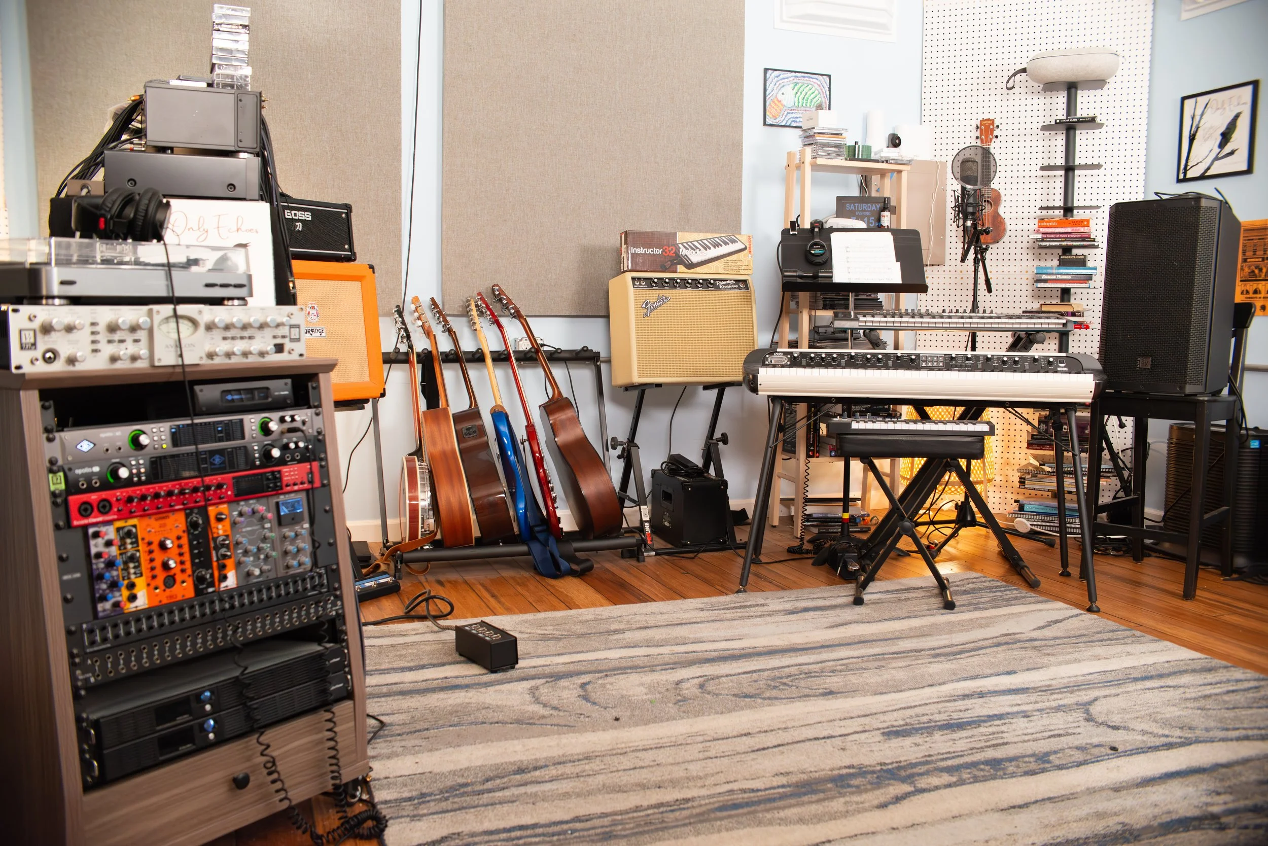 A music studio with various instruments and equipment, including guitars, keyboards, amplifiers, and audio gear, arranged in a cozy room with beige walls and a striped rug.