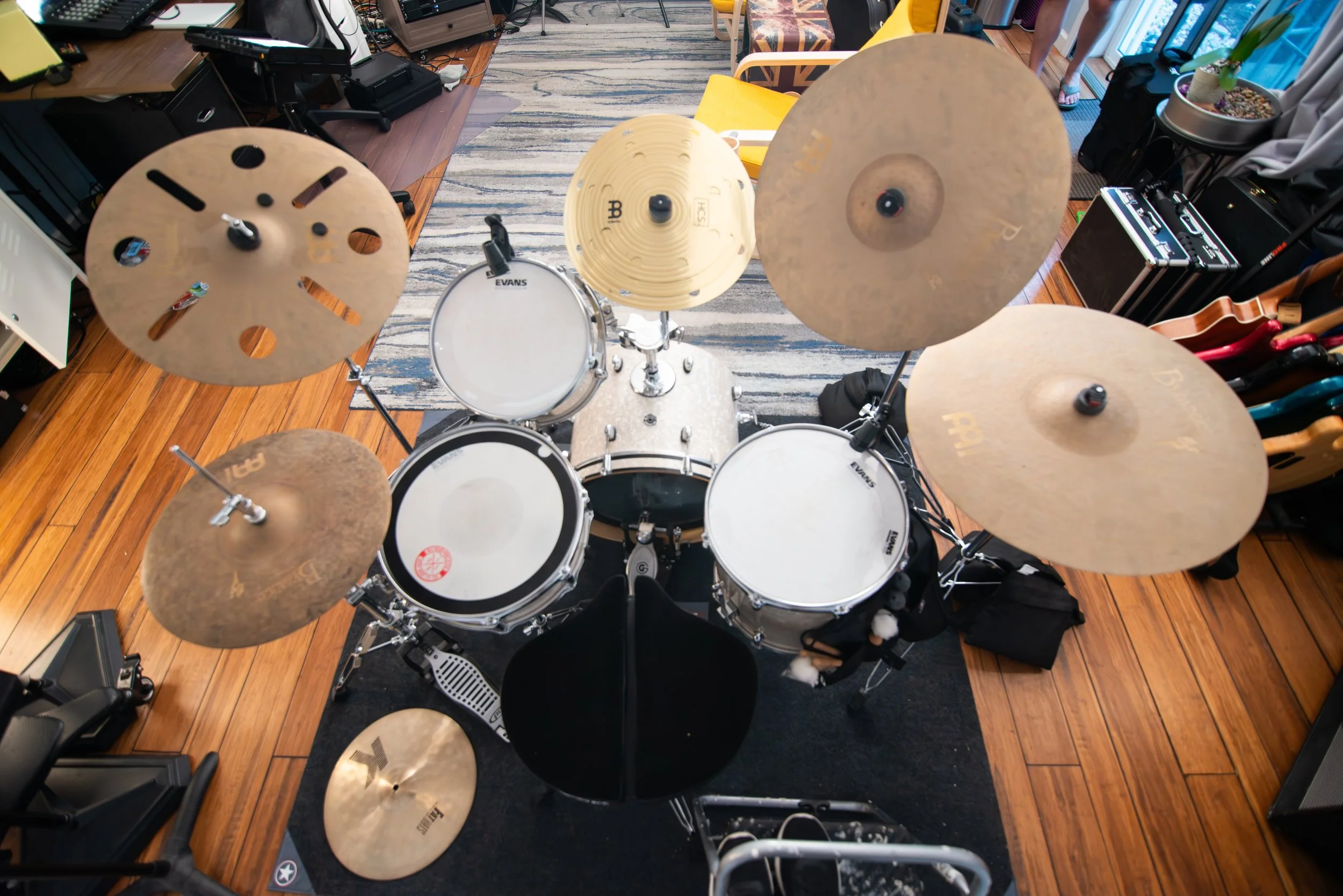 Top-down view of a drum kit with cymbals, surrounded by musical equipment and furniture in a room with wooden floor.