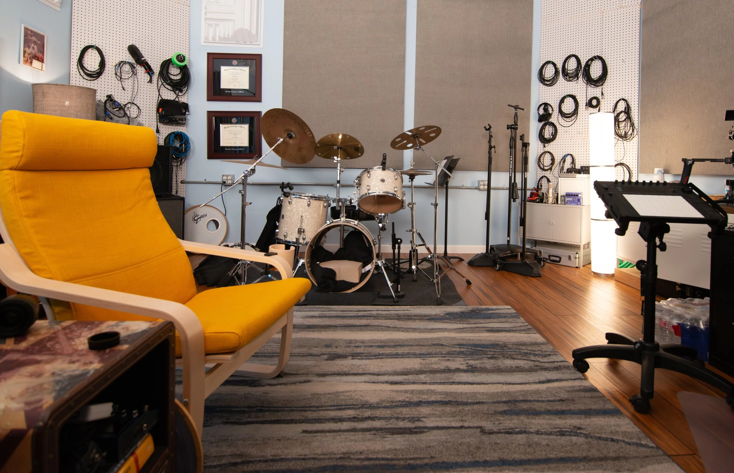 Music studio with a drum set, electronic keyboard, microphone stands, wall-mounted cables, framed diplomas, painting, and a yellow armchair on a striped rug.