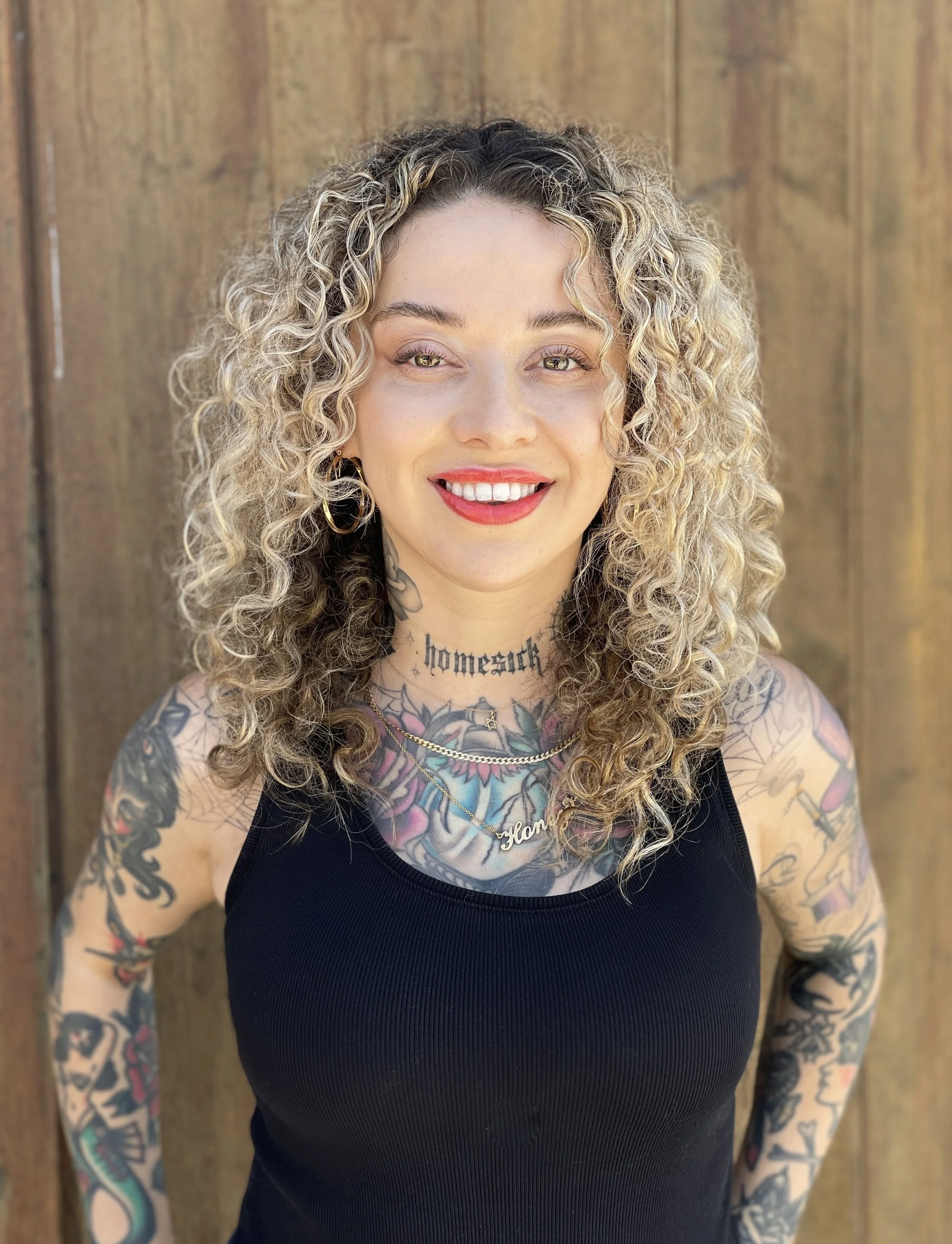 A woman with curly blonde hair, tattoos on her arms and chest, wearing a black tank top and gold jewelry, smiling outdoors against a wooden background.