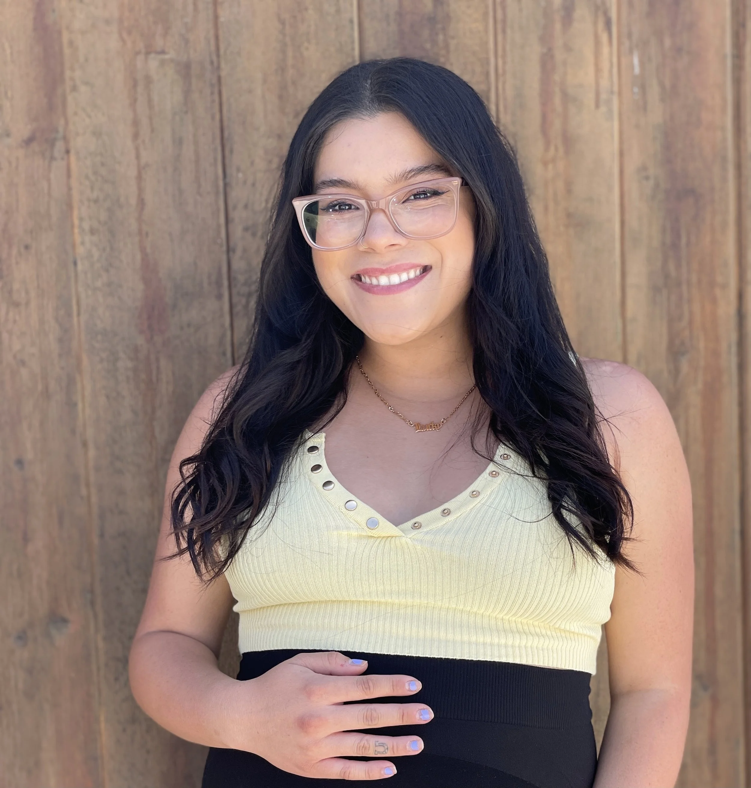 A young woman with long dark hair and glasses smiling, wearing a pale yellow top with metal buttons and a black bottom, standing in front of a wooden background.