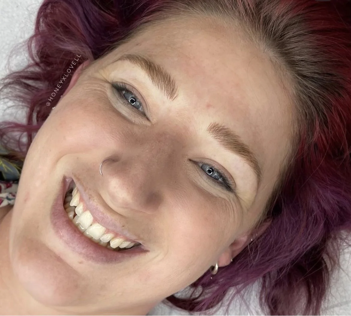 A young woman with pinkish-purple curly hair, light skin, blue eyes, and a nose piercing smiling widely, showing her white teeth.