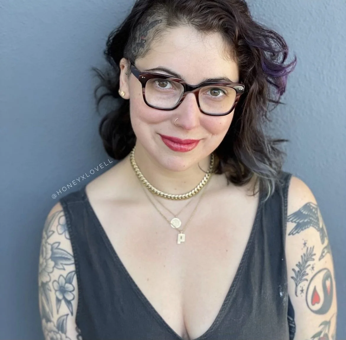 Portrait of a woman with glasses and dark hair with purple streaks, smiling, wearing a black sleeveless top and layered gold necklaces, tattoos on both arms, standing against a gray wall.