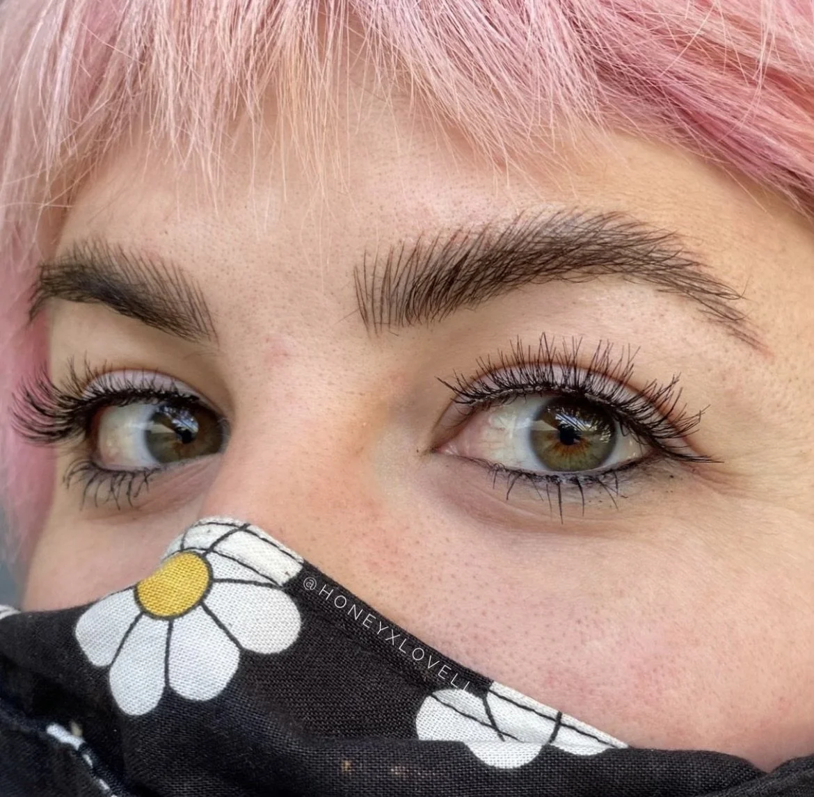Close-up of a person's eyes with pink hair, wearing mascara, and a face mask with daisy flower print covering their nose.