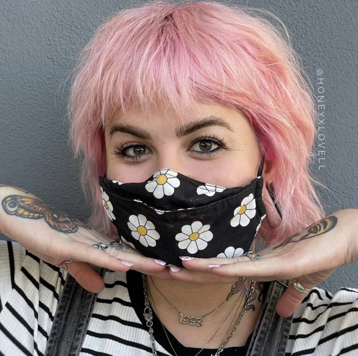 Young woman with pink hair wearing a black face mask with white daisies, making a gesture with her hands under her chin, standing against a gray background.