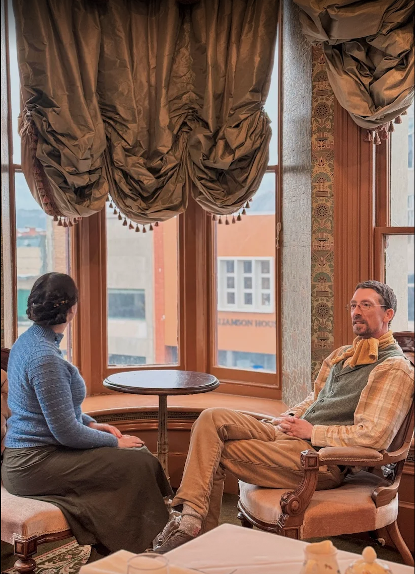 Two people sit facing each other in a room with large windows, heavy draped curtains, and a small round table between them, in a conversation.