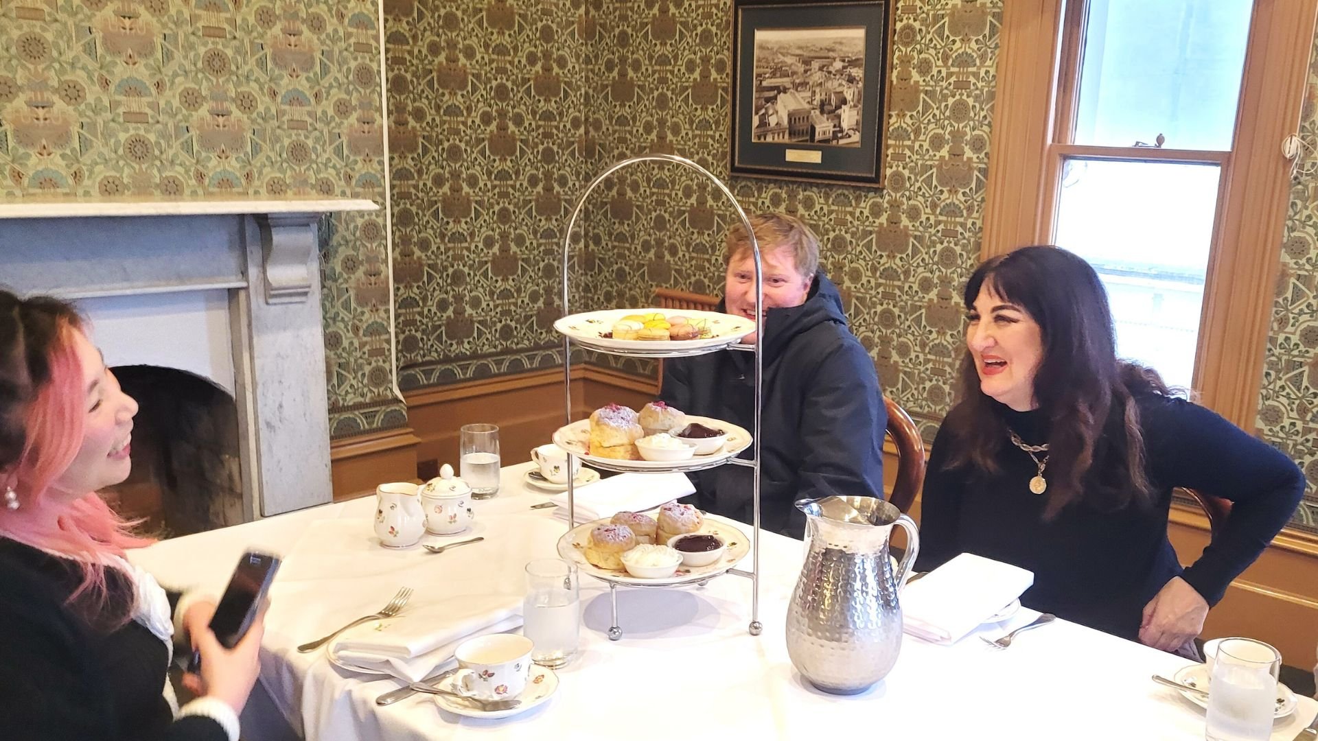 Three people enjoying an afternoon tea, with a three-tiered tray of assorted pastries and scones in the center of the table. The woman on the right is laughing, the man in the middle is smiling, and the woman on the left is holding a phone, all seate