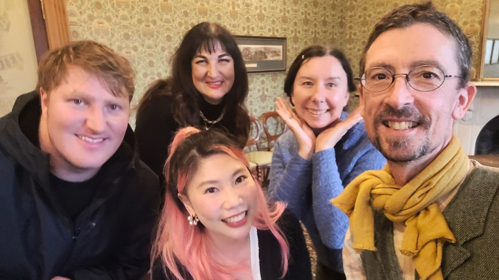 Group of six smiling people, three women and three men, gathered closely for a selfie in a cozy, vintage-style room.