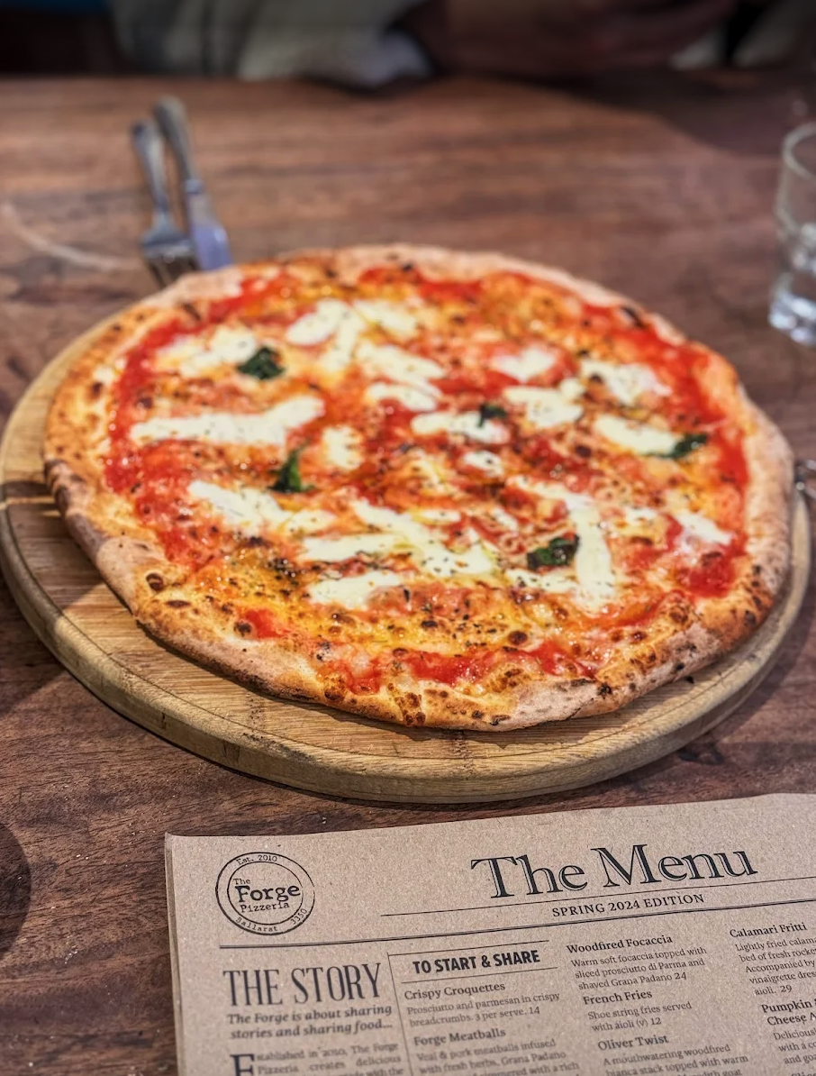 A pizza with mozzarella and tomato toppings on a wooden serving board on a wooden table, with a menu from The Forge Pizzeria in the foreground and a glass of water to the side.