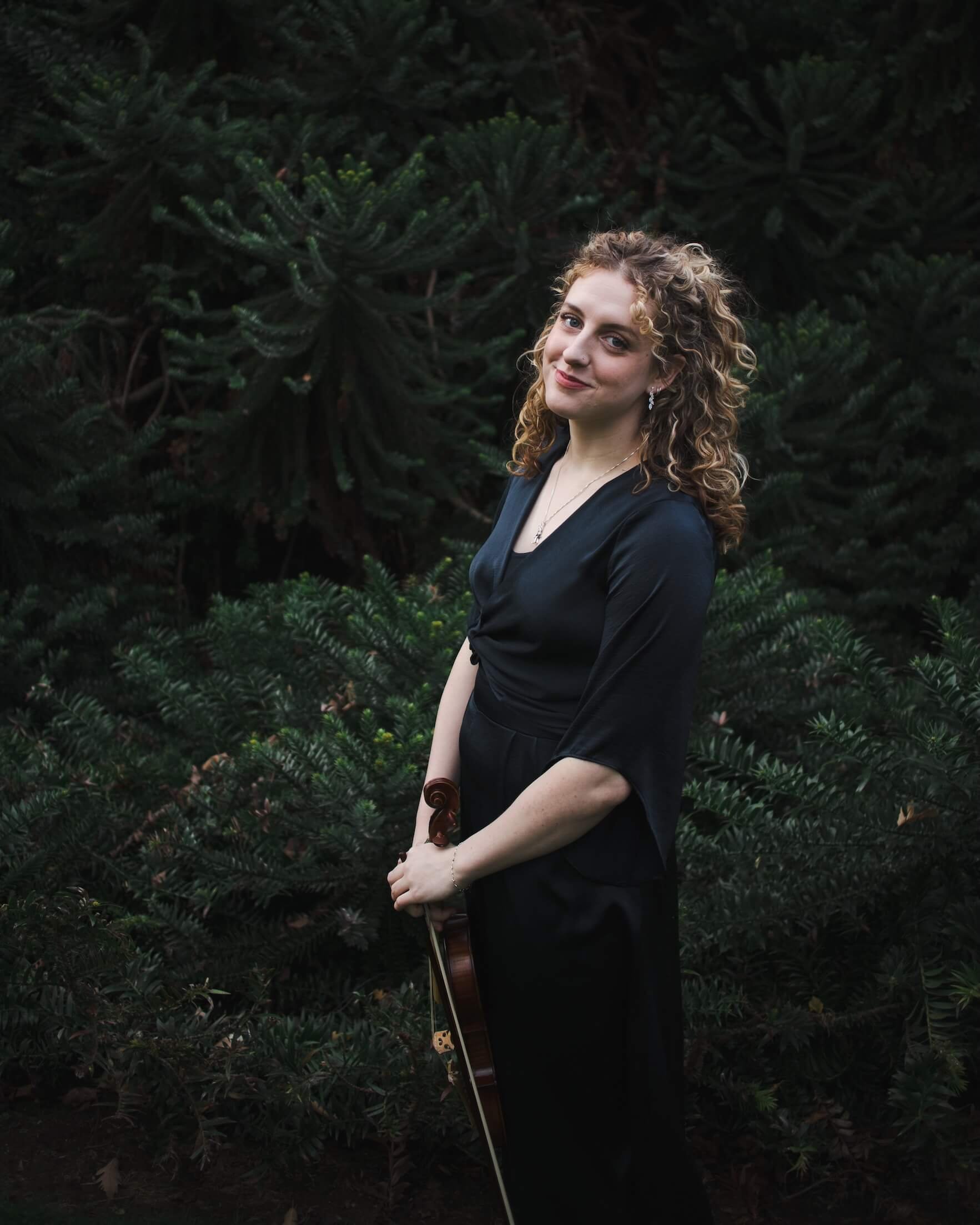 A woman with curly blonde hair and earrings standing outdoors against dark green foliage, holding a musical instrument in her hands.