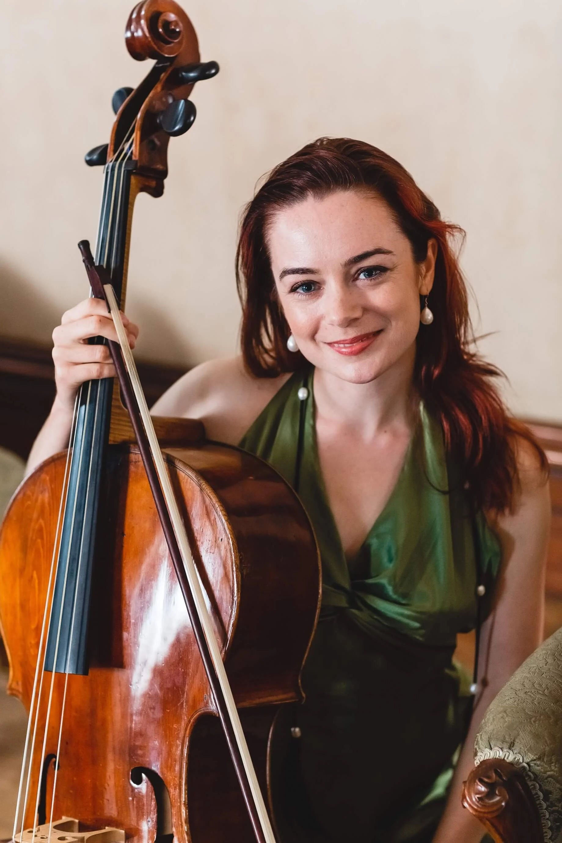 A woman with red hair and earrings smiling while holding a cello.