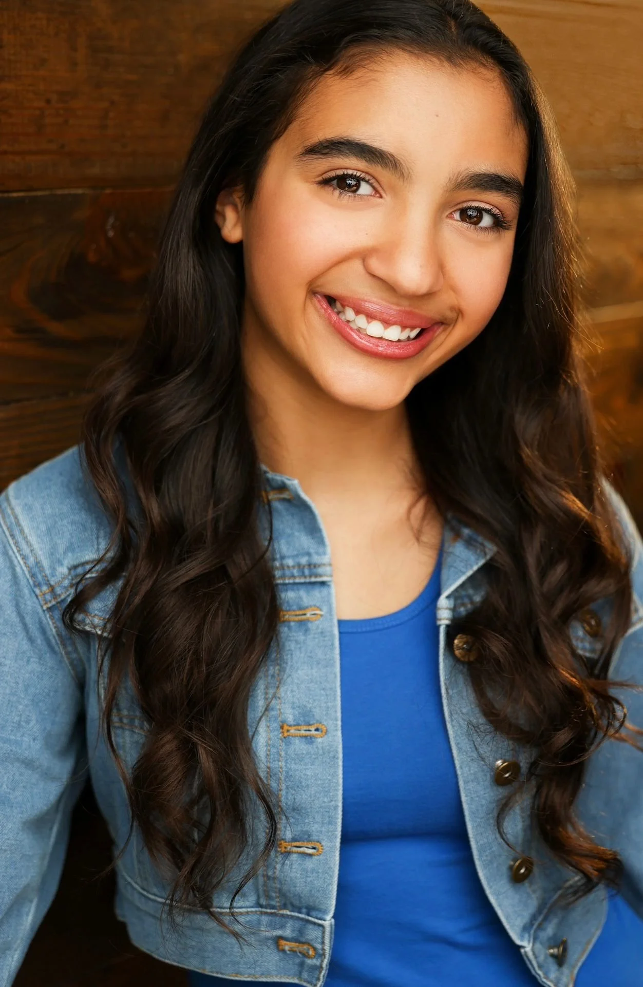 A young woman with long dark hair smiling, wearing a denim jacket and blue shirt, sitting in front of a wooden wall.
