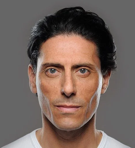 Close-up portrait of a man with dark hair, blue eyes, and a serious expression, wearing a white shirt against a gray background.