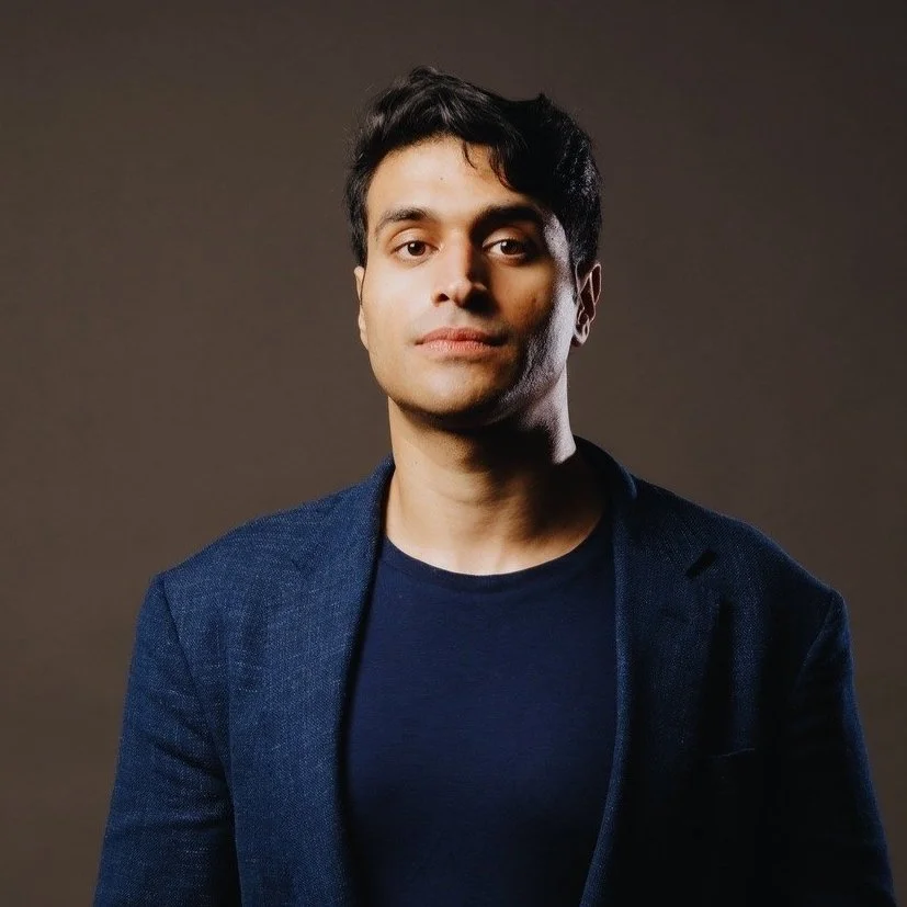 A young man with dark hair and light skin, wearing a navy blue blazer over a black T-shirt, standing against a dark brown background.