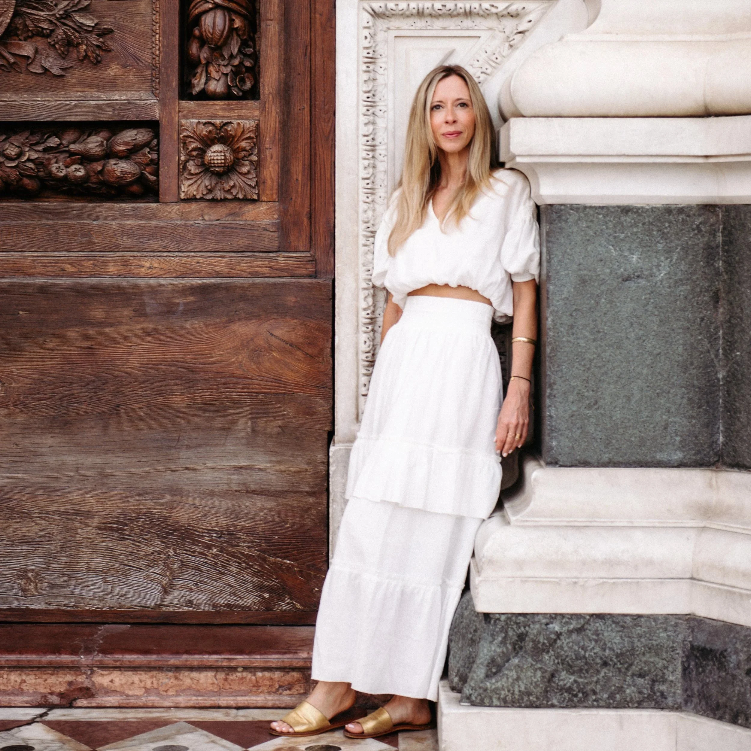 A woman with long blonde hair wearing a white pom-pom crop top, long white skirt, and gold sandals, leaning against a stone ledge beside a carved, dark wooden door with decorative floral carvings.