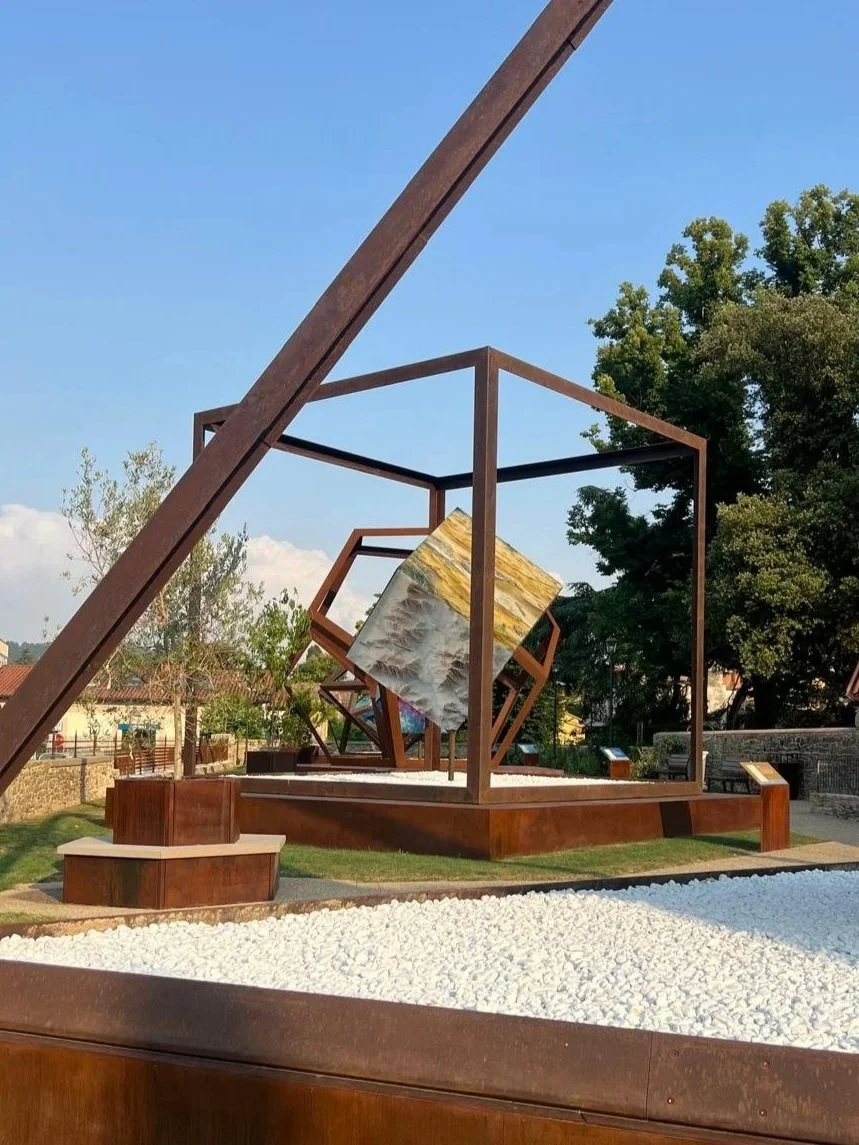 Escultura moderna de estructuras metálicas en forma de cubo y otros ángulos en un parque, con una pintura colgando en el centro y rodeada de grava blanca.