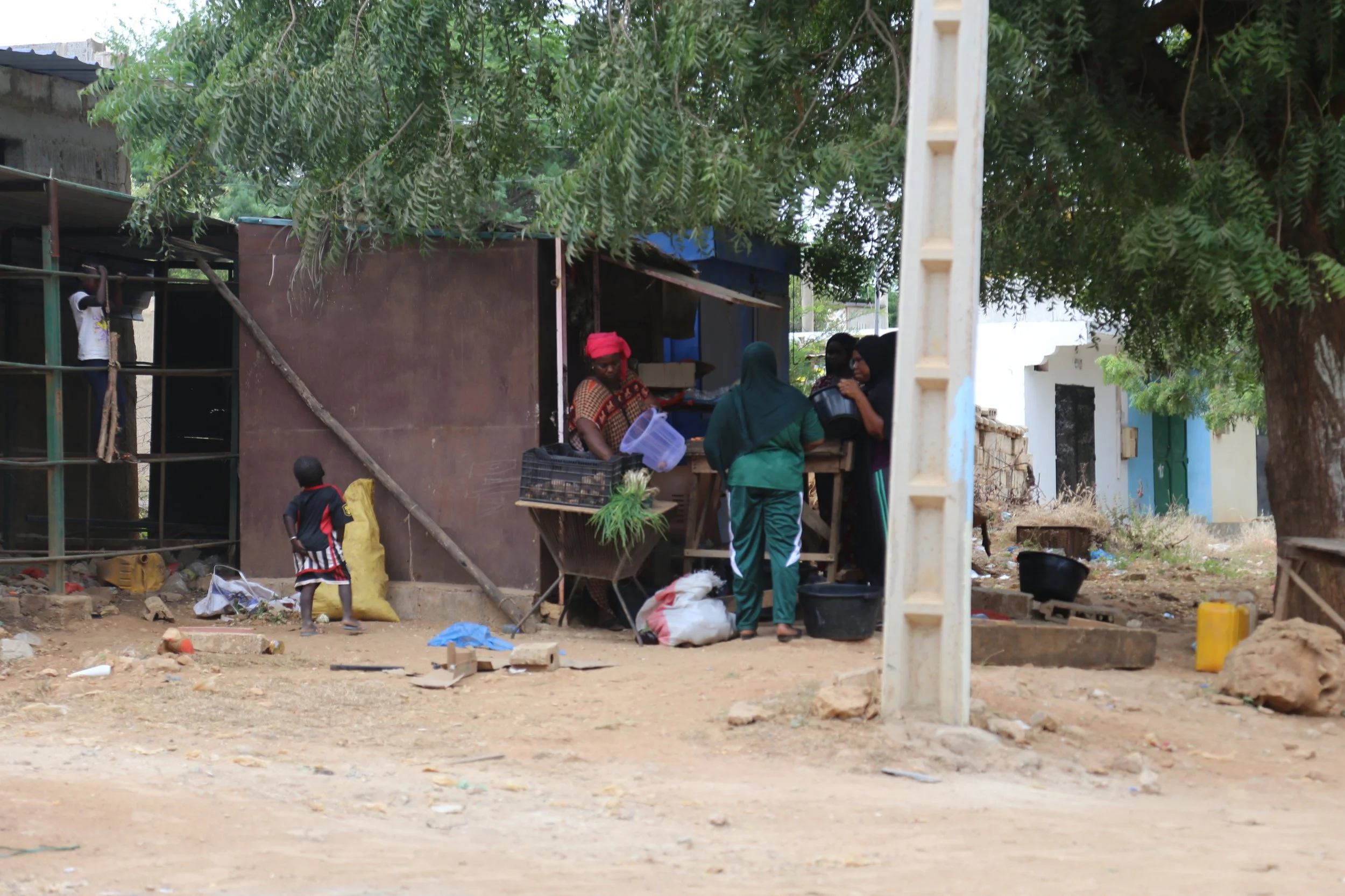 Personas en un mercado al aire libre, vendiendo y comprando productos, en un área con árboles y viviendas sencillas, piso de tierra y objetos diversos en el suelo.