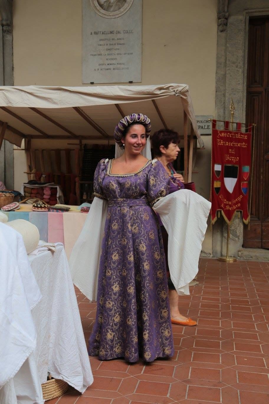 Mujer vestida con traje renacentista, de vestido largo y con tocado, en un mercado medieval, con otros puestos y una bandera en el fondo.