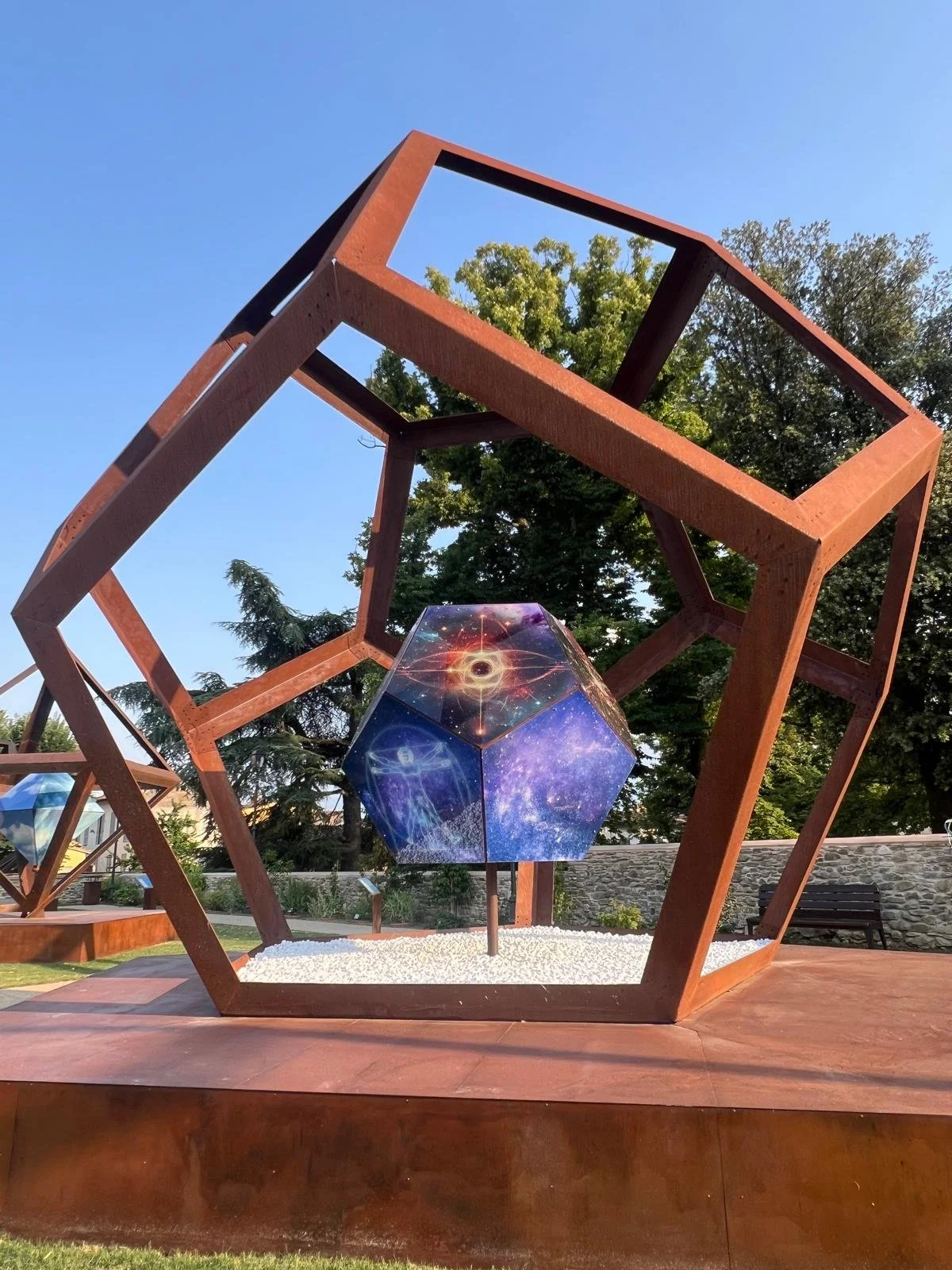 Escultura en un parque con estructura de metal y un dodecaedro decorado con imágenes del espacio, en un entorno con árboles y césped.