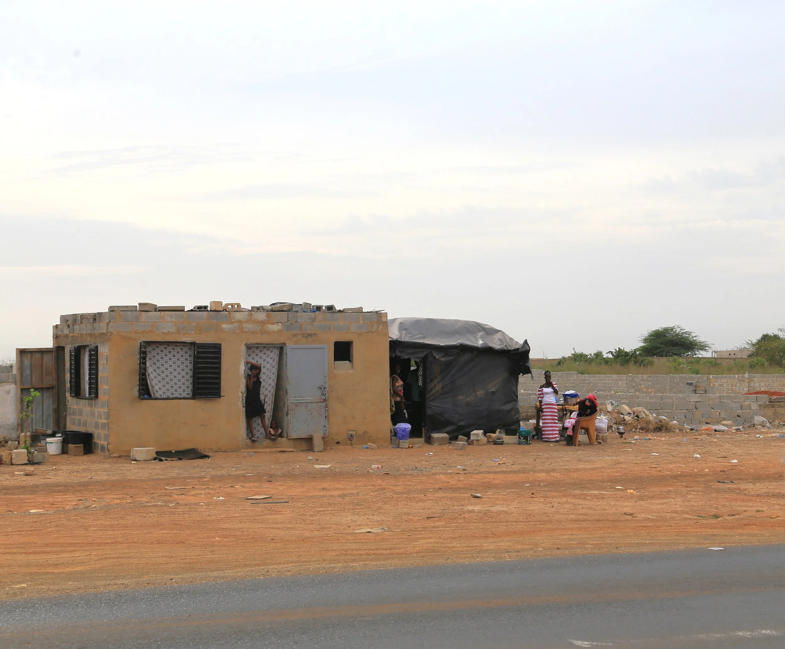 Casa sencilla en un entorno árido con personas reunidas afuera, algunas sentadas y otras de pie, en un día nublado.