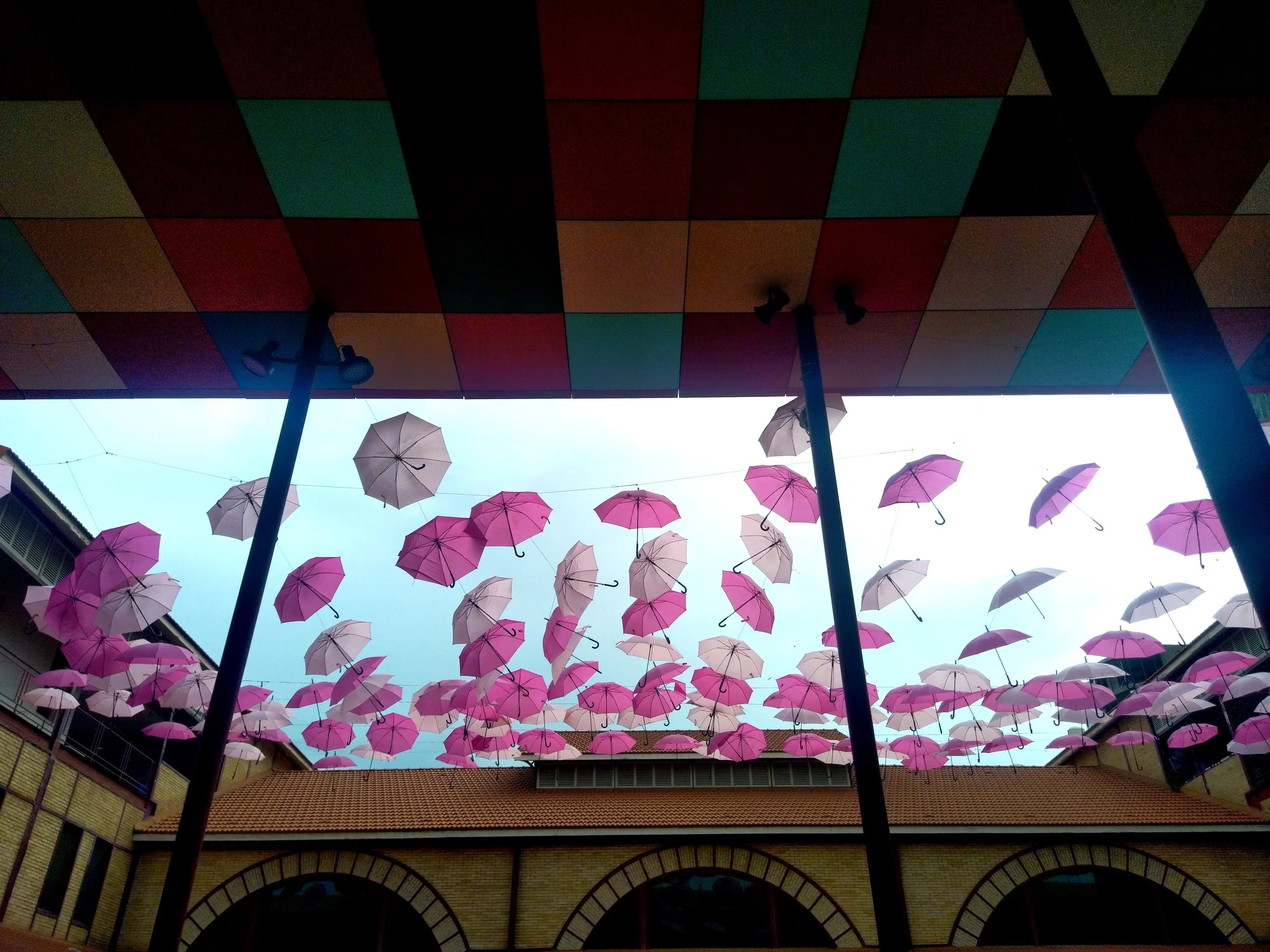 Techo con adoquines de colores y paraguas rosas y blancas suspendidos en el aire en un espacio abierto con edificios a los lados.