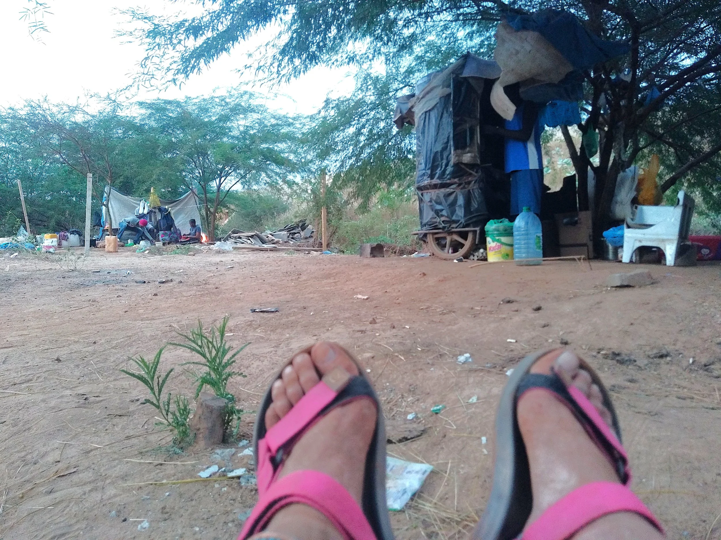 Vista desde el suelo de una persona con sandalias rosadas observando un campamento con dos tiendas de campaña y una comida en un entorno de tierra y árboles.