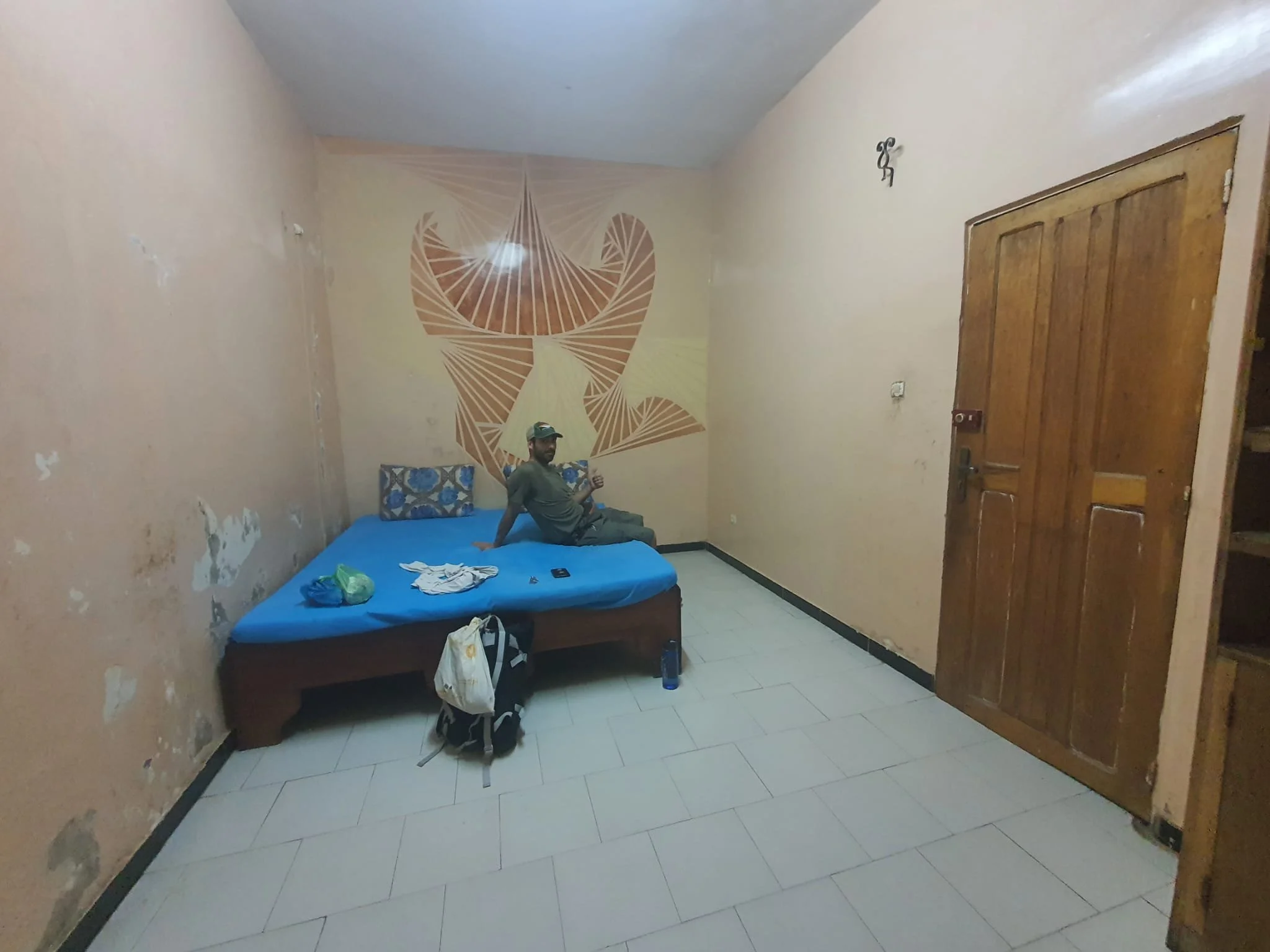 Hombre sentado en una cama en una habitación sencilla, con paredes de color beige y una pared decorada con arte en la cabecera. El suelo es de baldosas blancas y en el suelo hay una mochila, ropa, una botella y un control remoto.