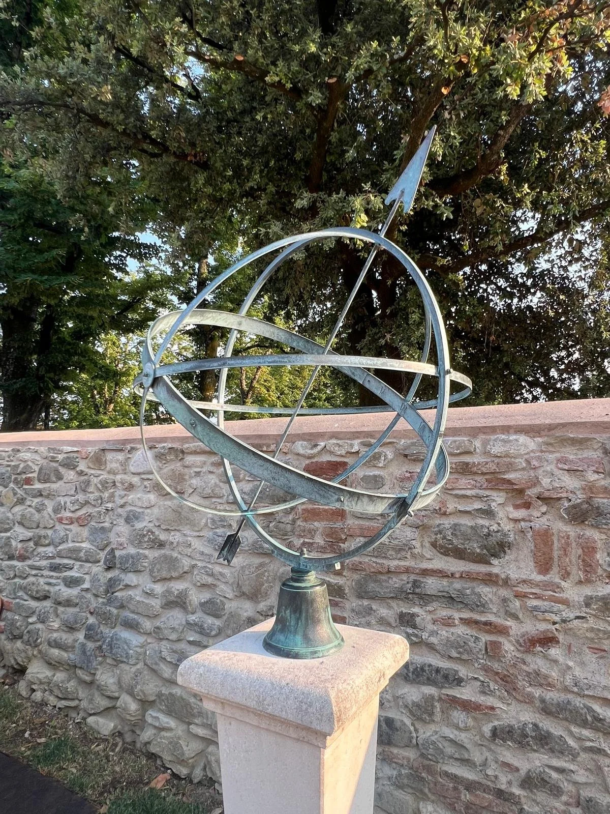 Escultura de un modelo astronómico de planetas y órbitas de metal en un pedestal de piedra junto a una pared de piedra y árboles en el fondo.