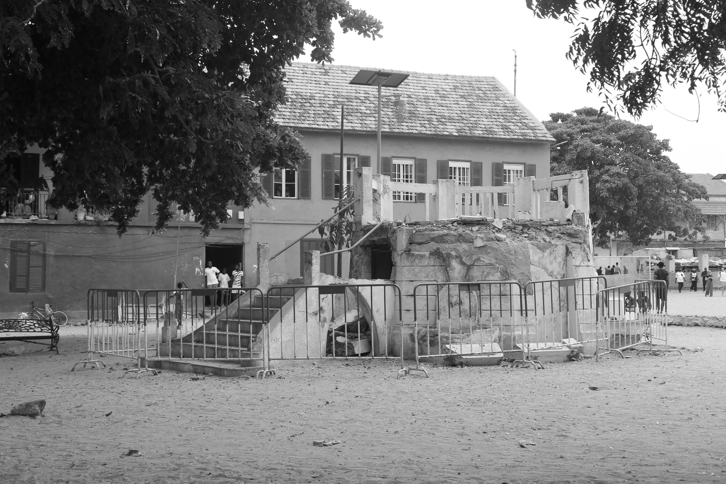 Una fuente de agua antigua en proceso de demolición en un parque, rodeada por una valla metálica, en un día nublado, con árboles y una casa en el fondo, y varias personas en la distancia.