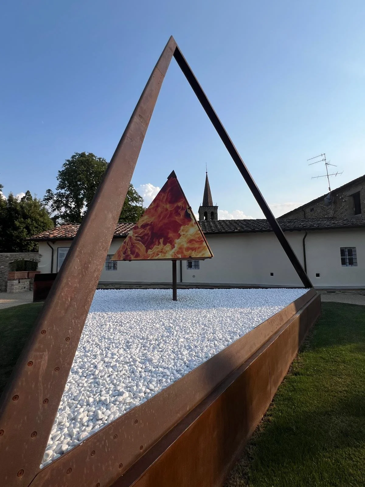 Escultura moderna en un parque con estructuras de metal en forma de triángulo, una de ellas con pintura de llamas, sobre base de piedras blancas. En el fondo, edificios antiguos y una iglesia con campanario, cielo despejado.