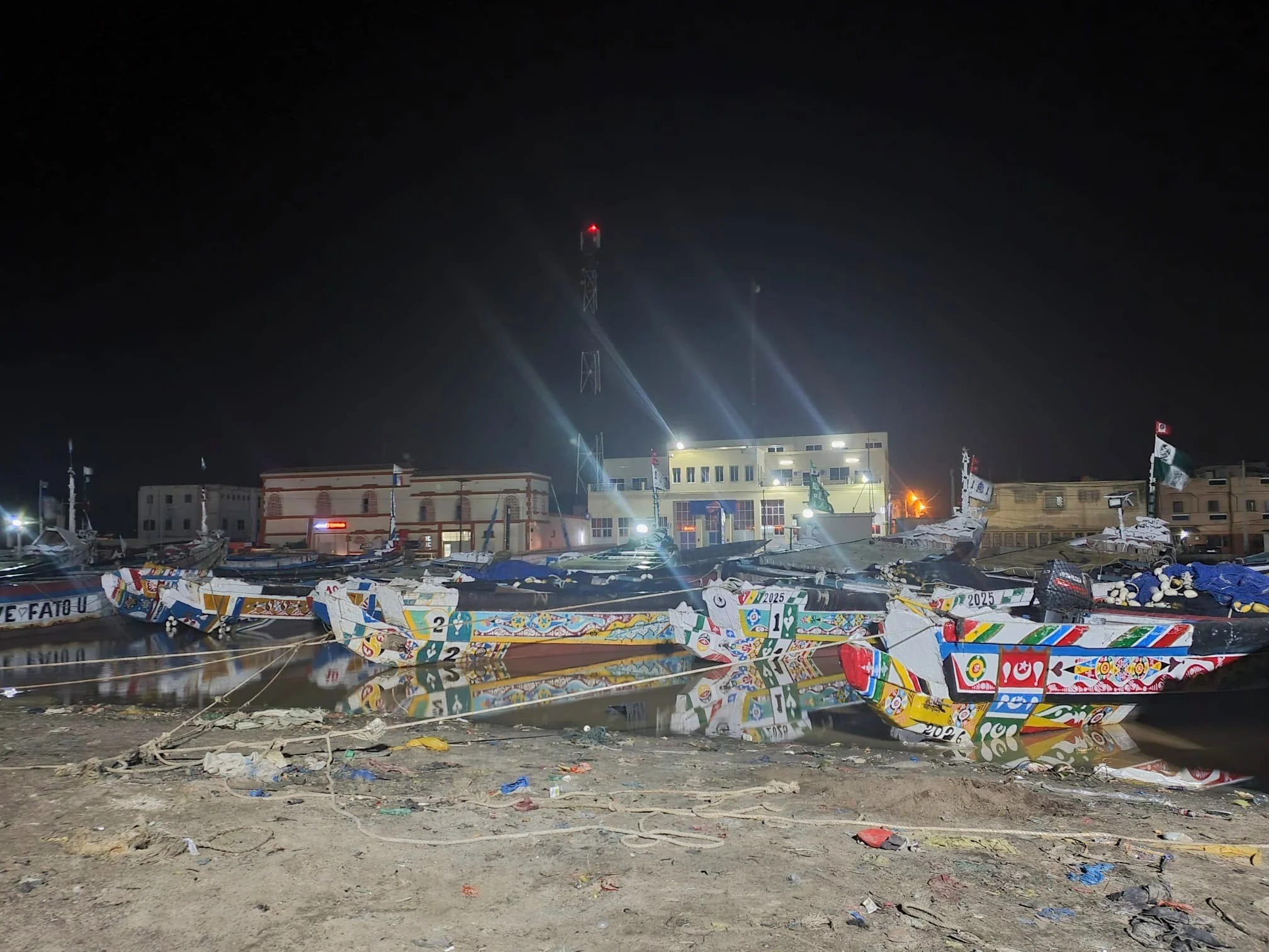 Barcos decorados con colores vibrantes y banderas en un muelle, reflejados en el agua, con edificios y un cielo nocturno en el fondo.