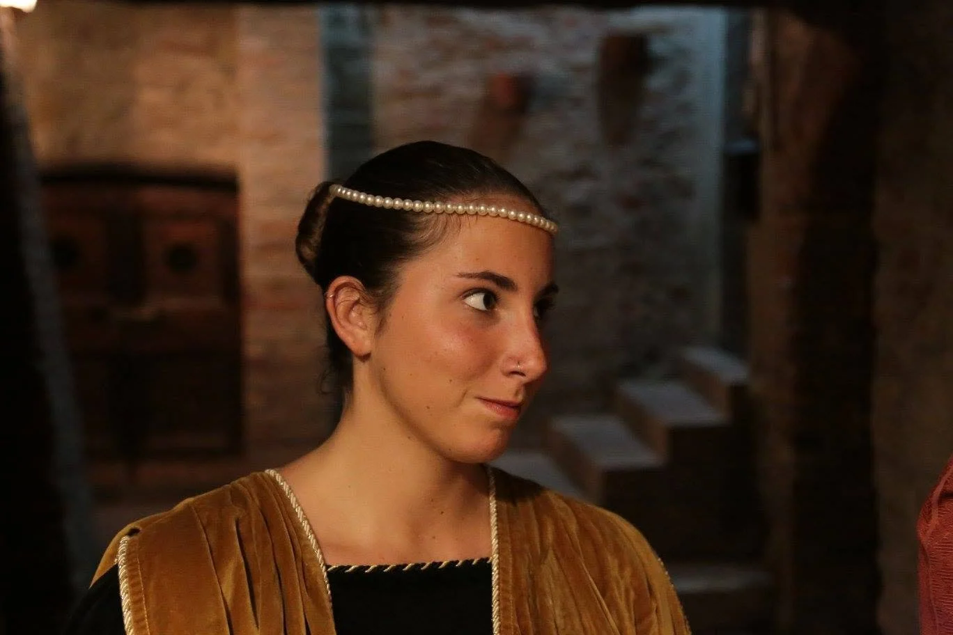 Mujer con pelo en un moño, usando una diadema de perlas, en un entorno rústico con paredes de ladrillo y escaleras de piedra.