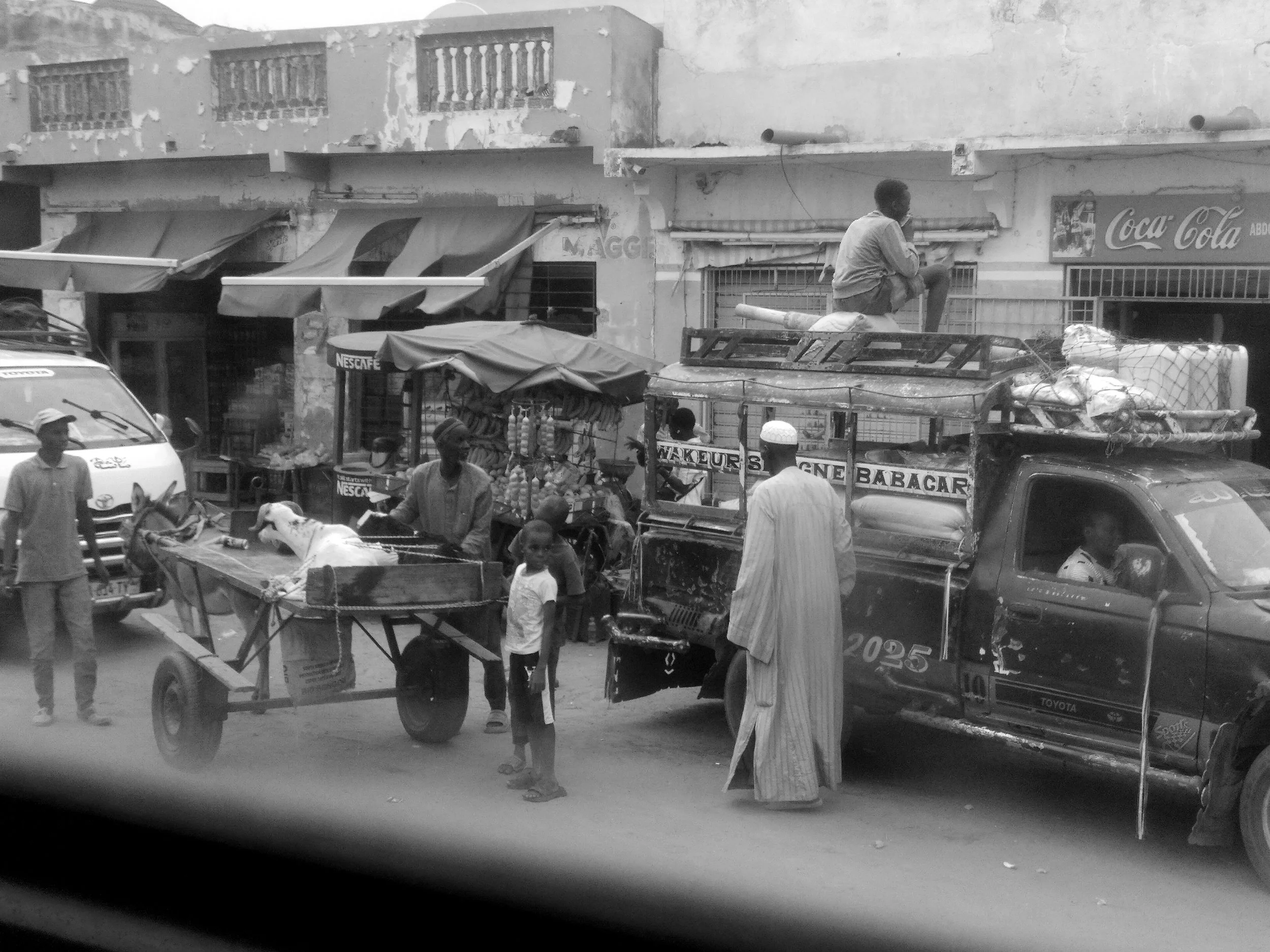 Escena urbana en una calle de una ciudad africana con personas, vehículos y puestos de venta callejera, en una foto en blanco y negro.