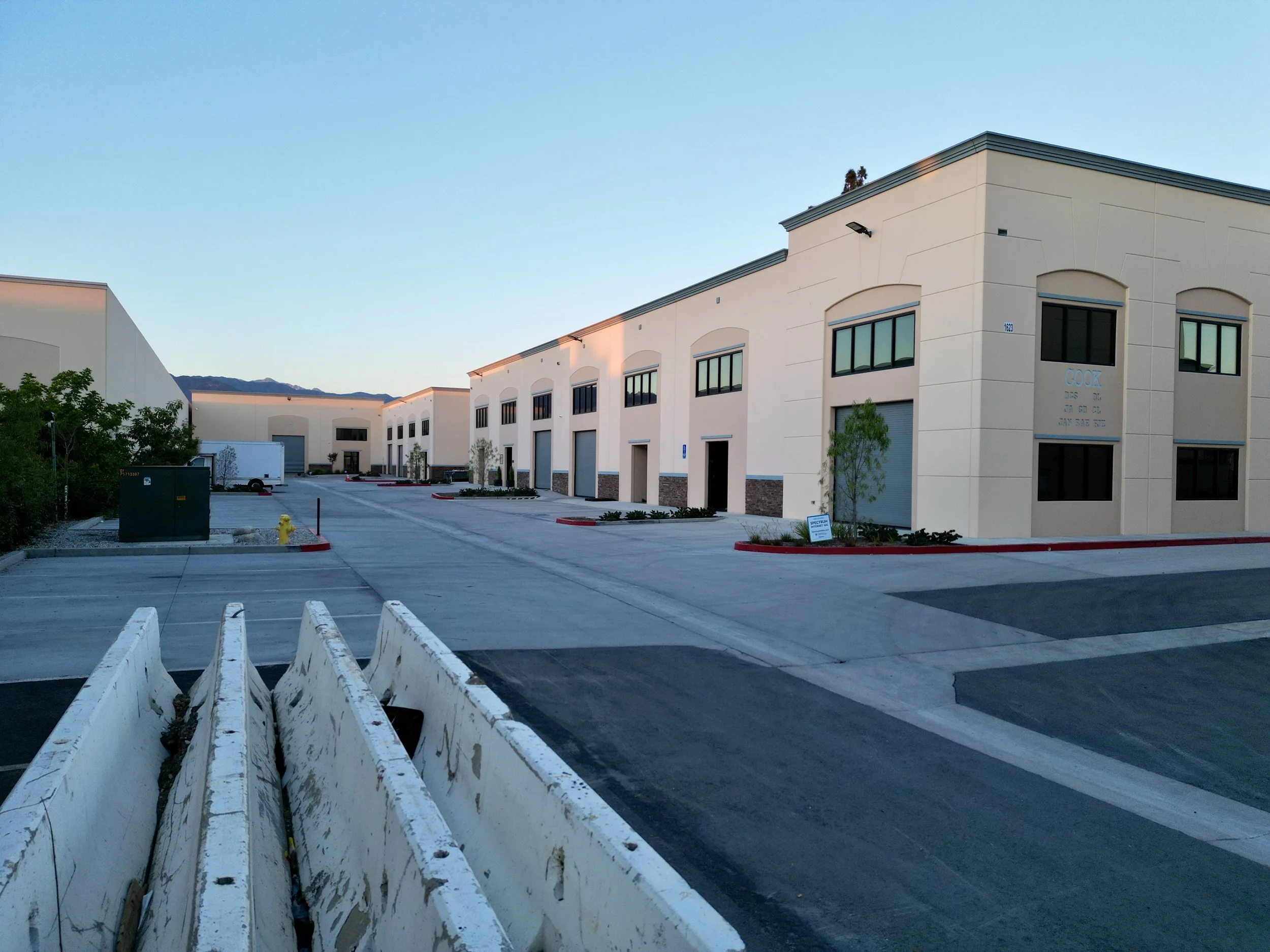 Empty industrial warehouse parking lot with tied-down construction barriers in the foreground, white warehouse buildings in the background, and mountains under a clear sky.